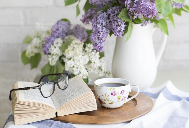 Kuvassa kirja, jonka päällä silmälasit, ja kahvikuppi pyöreän puutarjottimen päällä sekä taustalla kaksi kukkamaljakkoa, joissa sini-valkoiset kukat