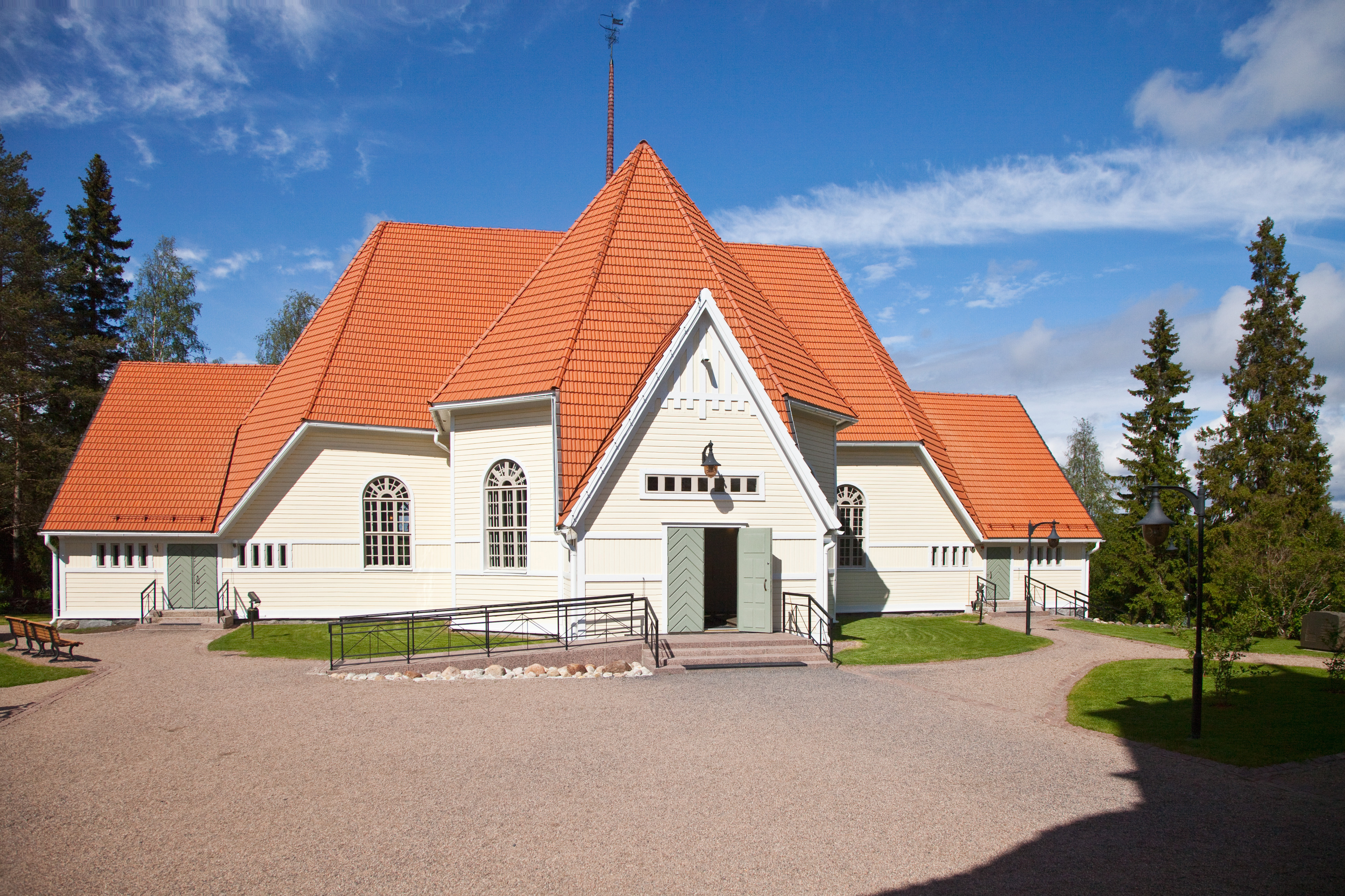 Kuvassa on kesäinen Haukiputaan kirkko ulkoa kuvattuna. Punainen katto hohtaa auringossa.