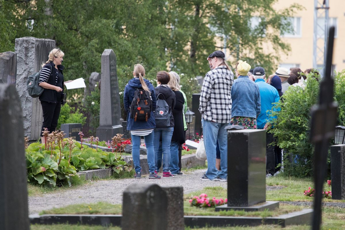 Ryhmä ihmisiä seisoo hautausmaalla ja kuuntelee oppaan puhetta.