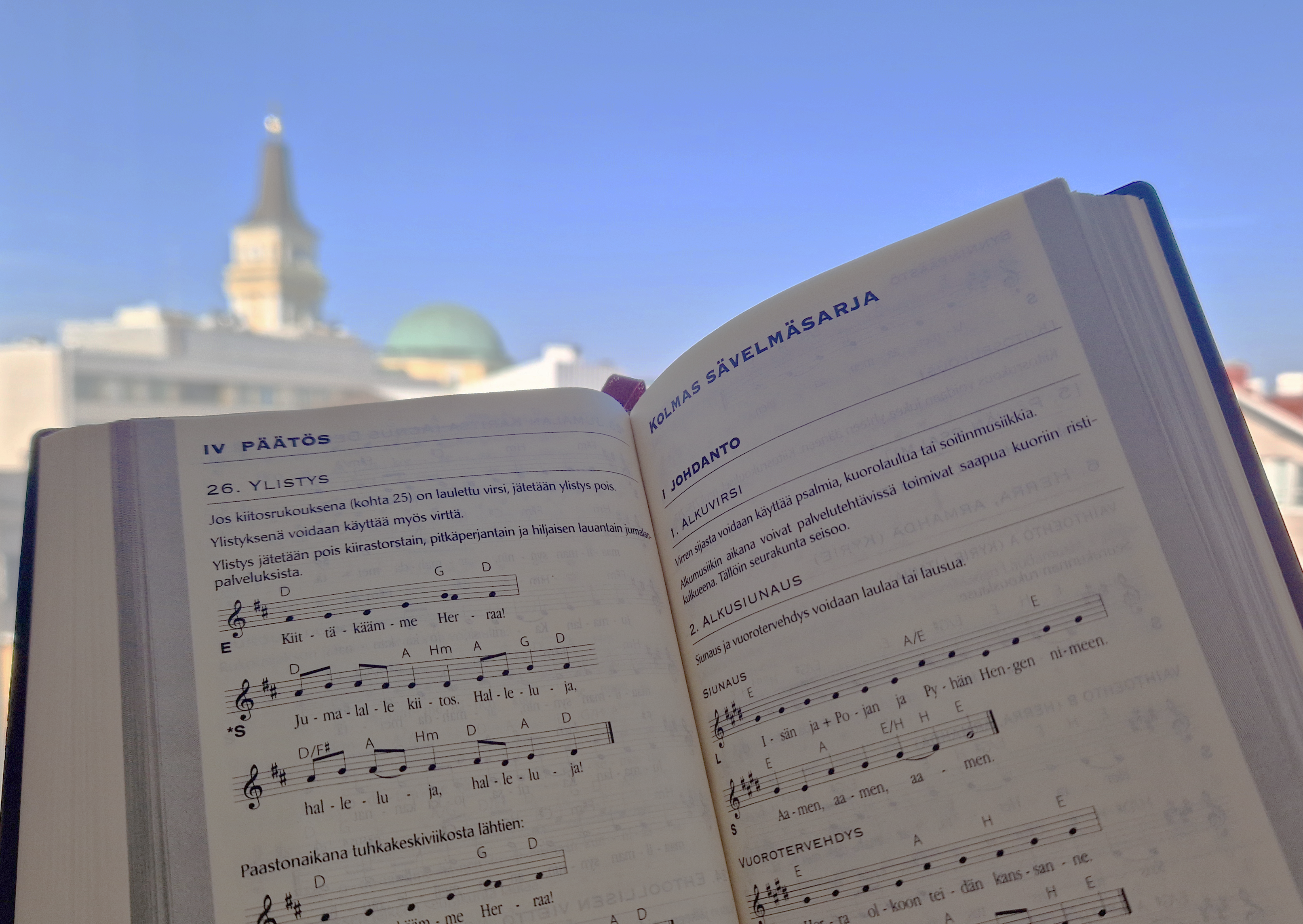 Taustalla on tuomiokirkko ja edessä virsikirja avoinna kolmannen sävelmäsarjan kohdalta
