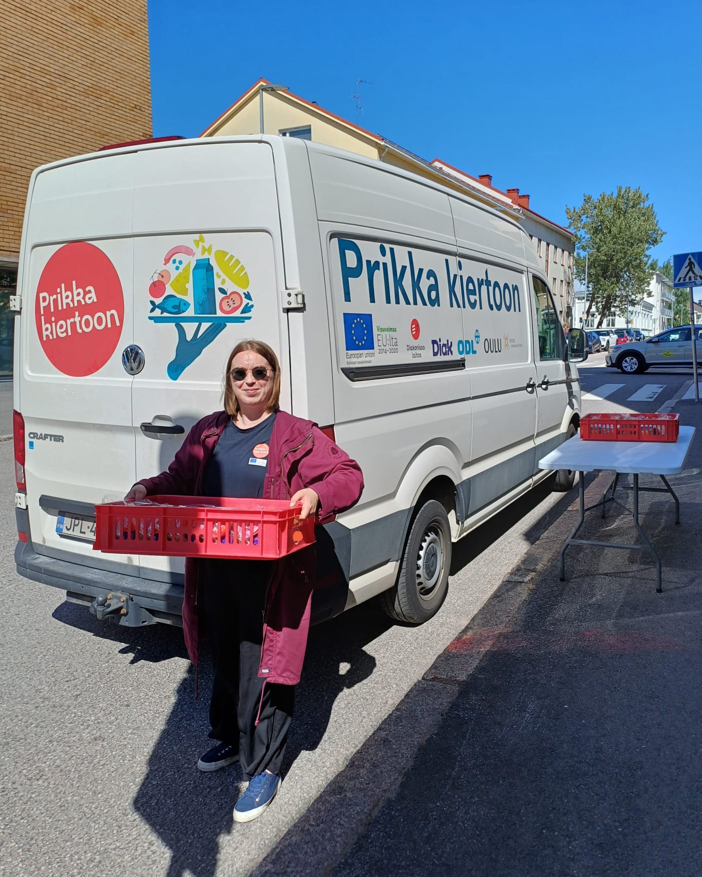 Prikka kiertoon -hankkeen logoilla koristeltu pakettiauto, jonka vieressä seisoo nuori nainen ruokakori käs...