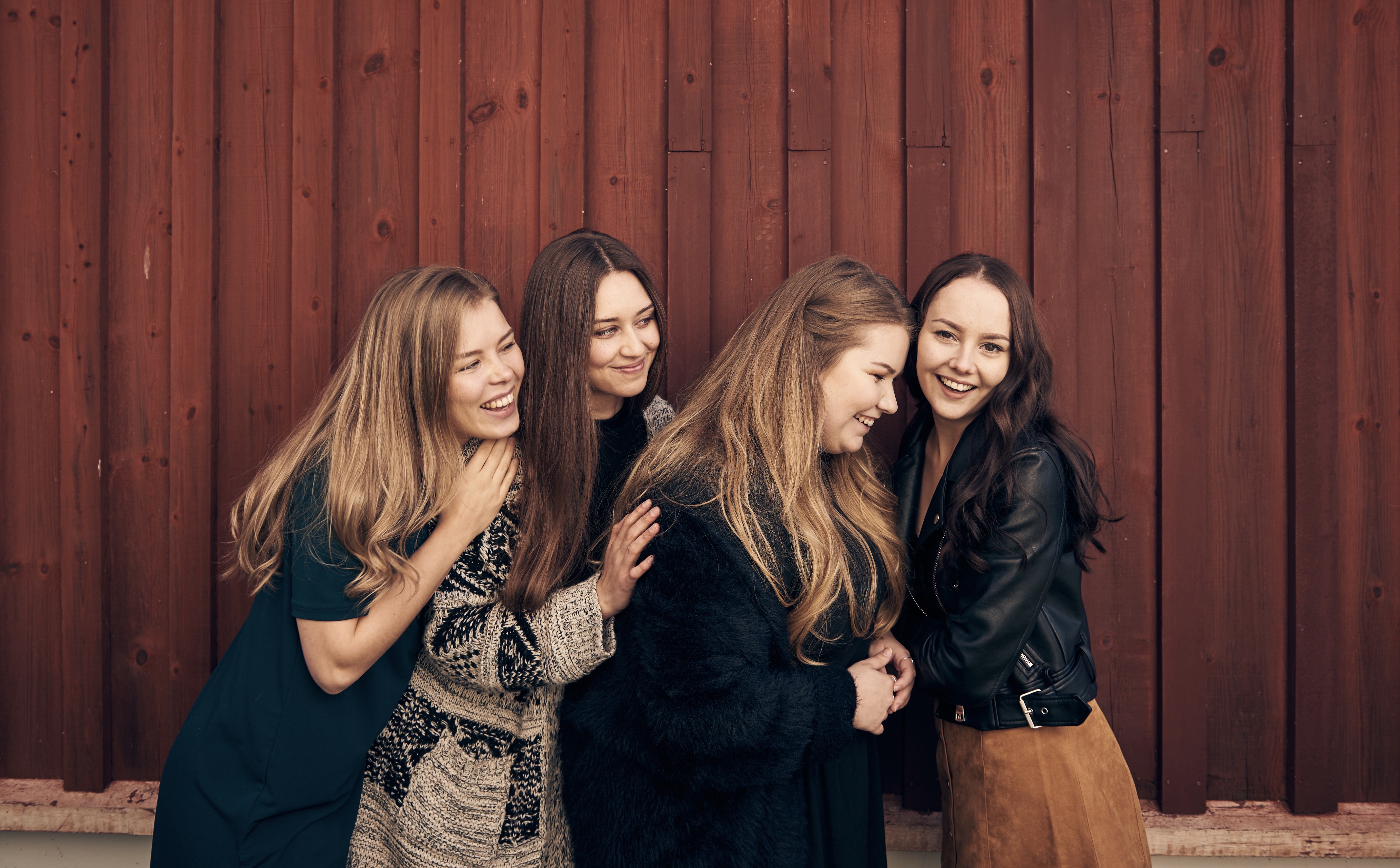 Kuvassa neljä nuorta iloista naista hymyilee punaisen puutalon edustalla. He ovat lauluyhtye Neliä.