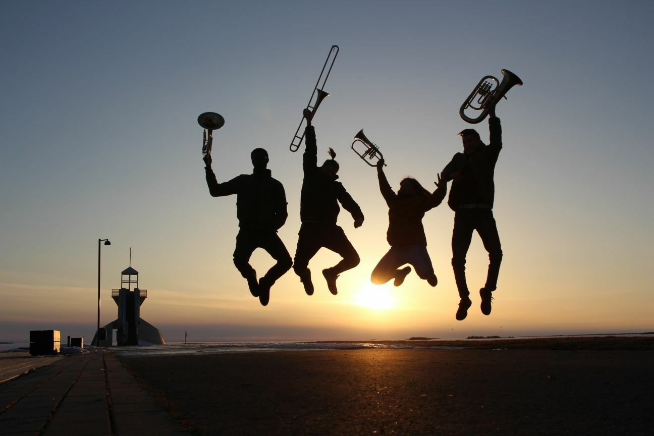 Neljä soittajaa hyppää ilmaan soittimet käsissään (käyrätorvi, pasuuna, trumpetti ja baritonitorvi). Kuva Kimmo Pihlajamaa.
