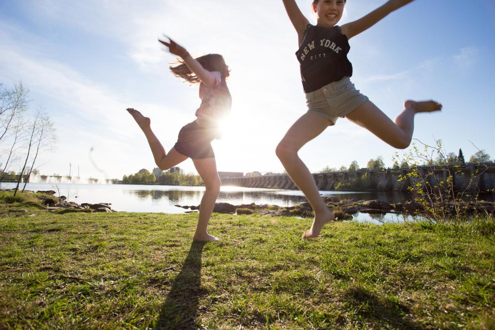 Kaksi nuorta tyttöä hyppää kesäisessä maisemassa.
