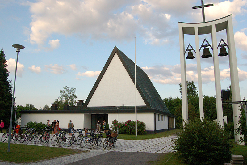 Valkoinen kirkkorakennus ulkoa päin, edessä muutama ihminen pihalla ja pyöriä