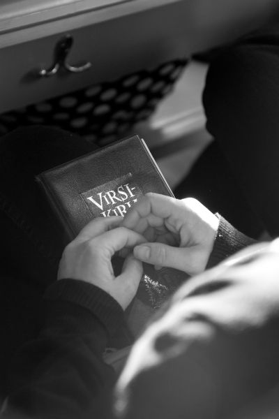 Mustavalkoinen valokuva, ihminen istuu kädet ristissä kirkon penkissä, virsikirja sylissään. Kuva: Sanna Kr...