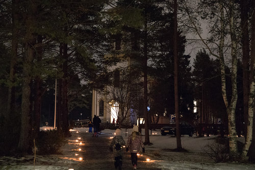 Kuvassa on ulkotulin valaistu kävelytie, jolla kulkee ihmisiä kohti kirkkoa.