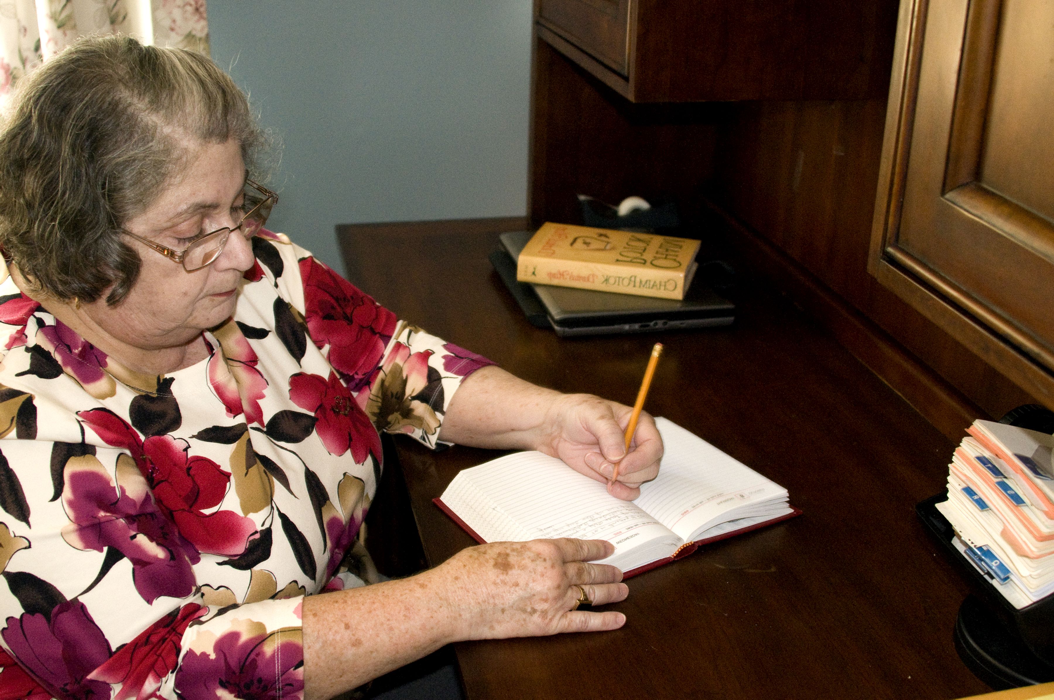 Iäkäs nainen istuu pöydän ääressä ja kirjoittaa paperille.