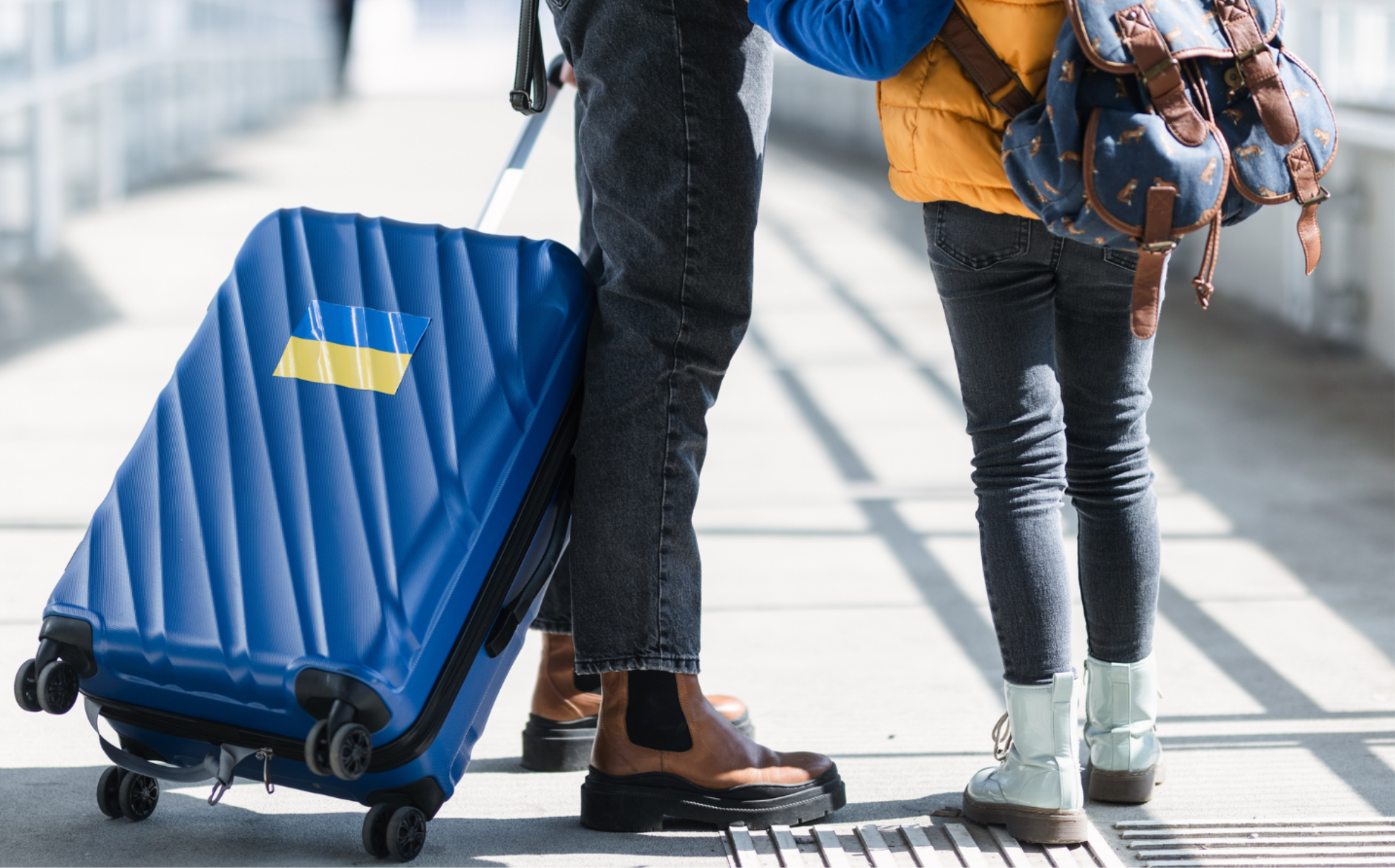 Kaksi ihmistä, joista toisella on reppu selässä ja toinen vetää sinistä matkalaukkua, jossa on Ukrainan lippu.