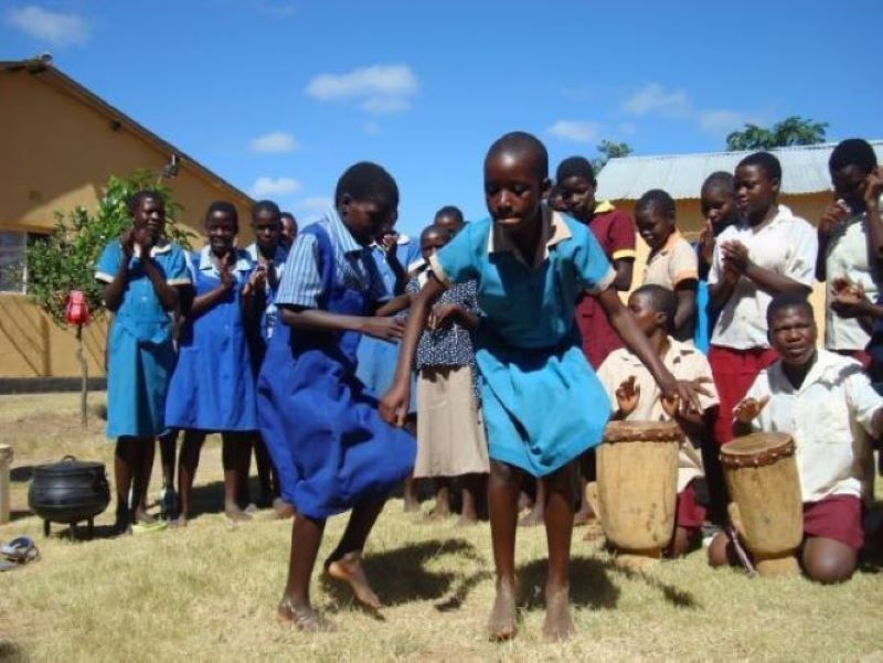 Kuvassa tummaihoiset lapset tanssivat etualalla sinisissä koulupuvuissaan ja taustalla toiset lapset taputtavat ja soittavat rumpua 