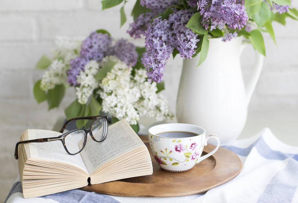sinivalkoisia kukkia taustalla, edessä avoin kirja, jonka päällä silmälasit ja vieressä kahvikuppi, jossa kukkien kuvia